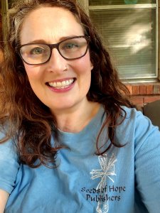 A middle-aged Caucasian woman with long dark brown hair and glasses is smiling at the camera. She is a wearing a light blue T-shirt with a logo on her right shoulder. It is a customized cross with a shaft of wheat. The words "Seeds of Hope Publishers" is written across it. 