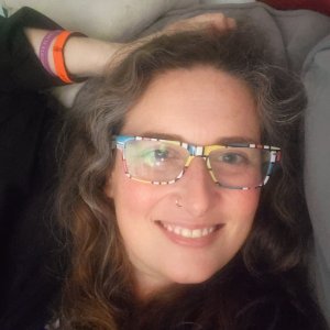 Close-up headshot of a middle-aged woman leaninng back on her couch. Her hand is relaxed on her head and she is smiling at the camera. She is wearing eyeglasses that are blocks of yellow, red. blue and white in design. She is wearing two bracelets on the arm resting on her head, One is orange and the other is purple. The woman pictured is Dawn Michelle Michals.