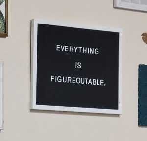 A white-framed picture hanging on a light beige wall. The picture is all black with the words "everything is figureoutable." written in white in all caps. 